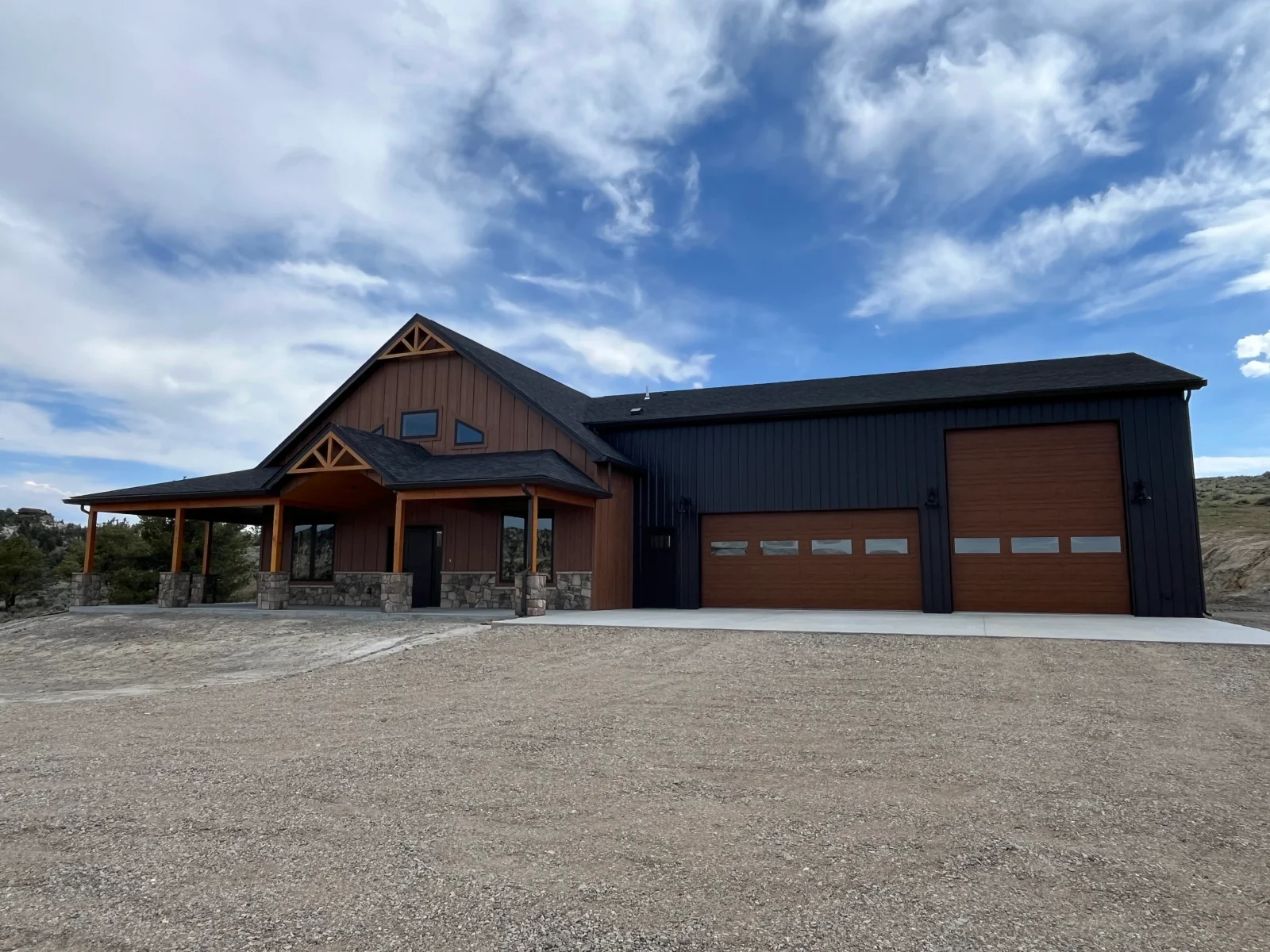 Modern home with brown metal siding, stone foundation, timber gable details, and attached black garage with metal doors