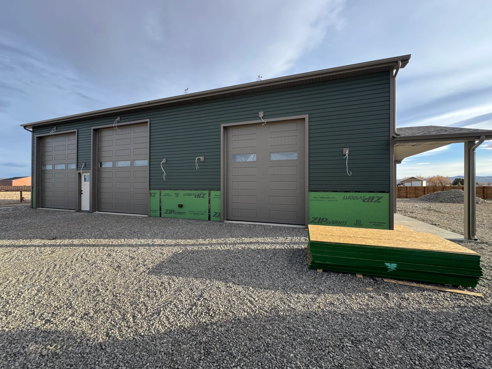 Three-bay garage with sage green siding, gray overhead doors, and attached carport structure