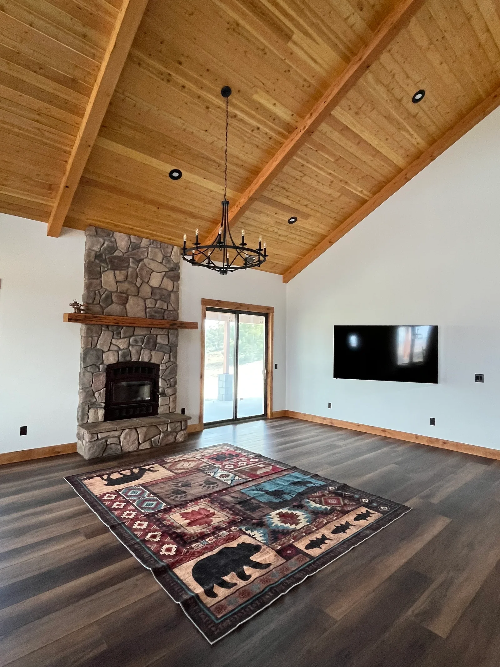Open great room with exposed wood beams, stacked stone fireplace, hardwood floors, and black chandelier